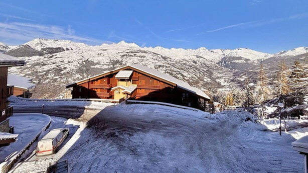 A wooden chalet with a snow-covered roof, set against a backdrop of snow-capped mountains under a clear blue sky.