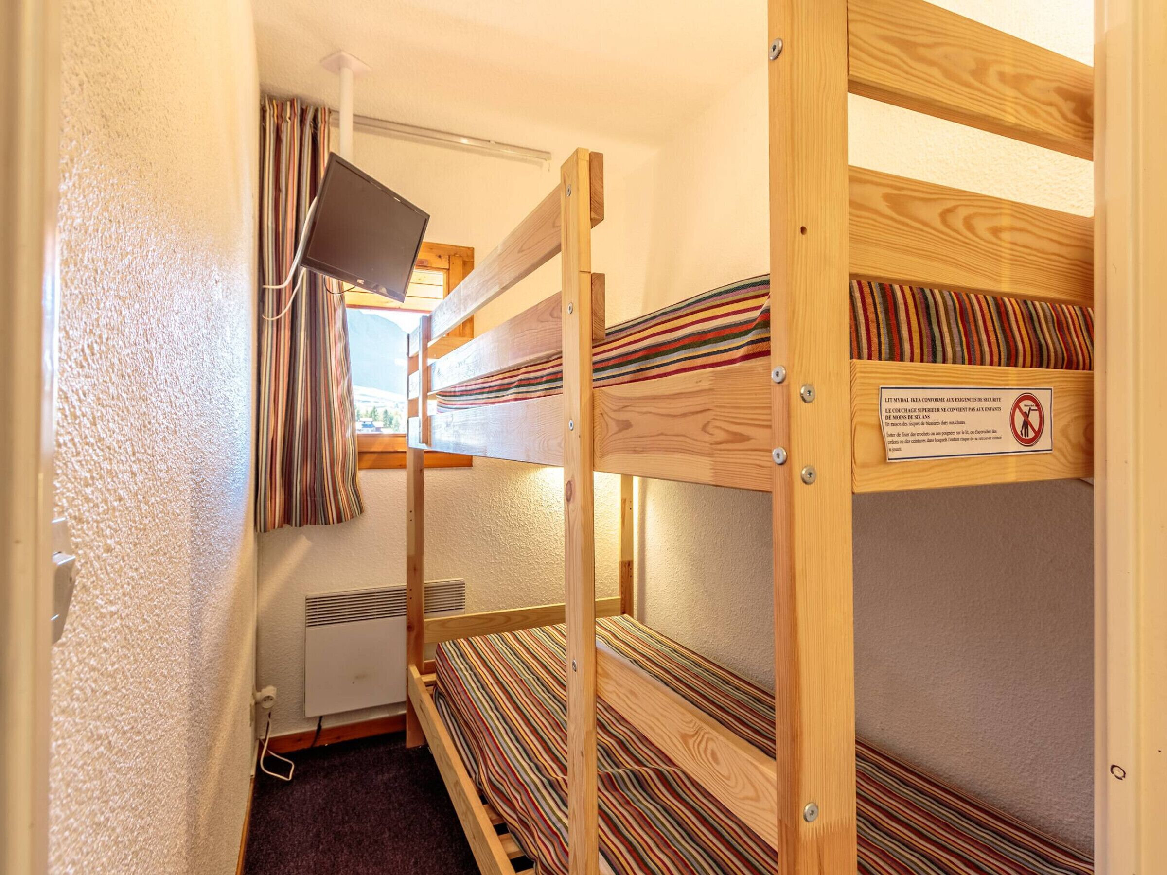 Quarto com beliche, uma televisão no beliche superior, janela com cortinas e um aviso no beliche inferior.