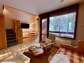 A cozy room with a wooden staircase, a dining area, a sofa, a rocking chair, and a large window with a view of snow-covered trees. - Duplex 6 pers, central location, parking, pets OK (La Plagne)