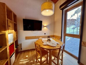 A cozy room with a wooden dining table, chairs, a television mounted on the wall, and a large window offering a view of a snowy landscape.