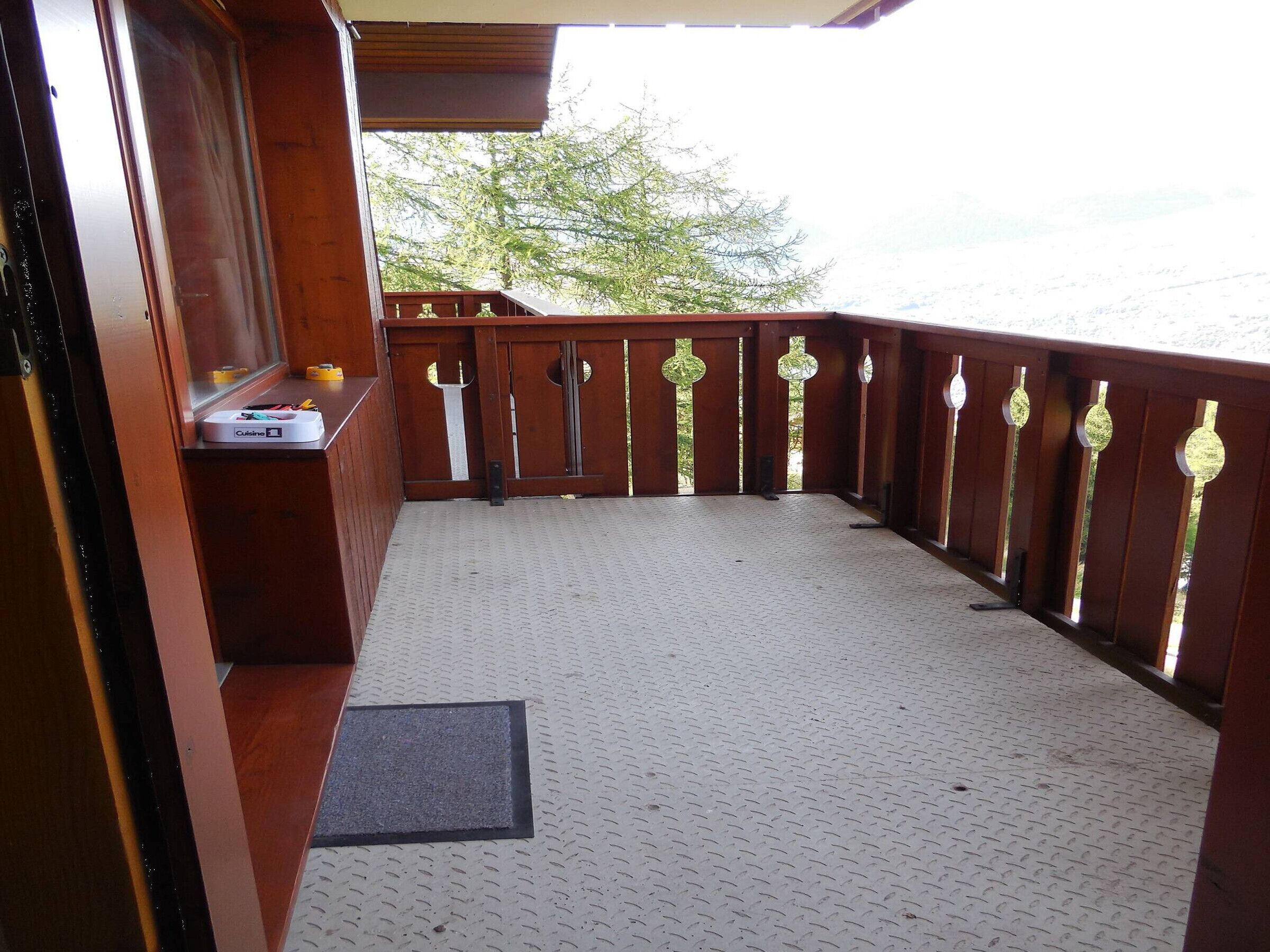 Un balcon avec une balustrade en bois, une petite table sur laquelle se trouve un appareil, et un paillasson sur un sol texturé.