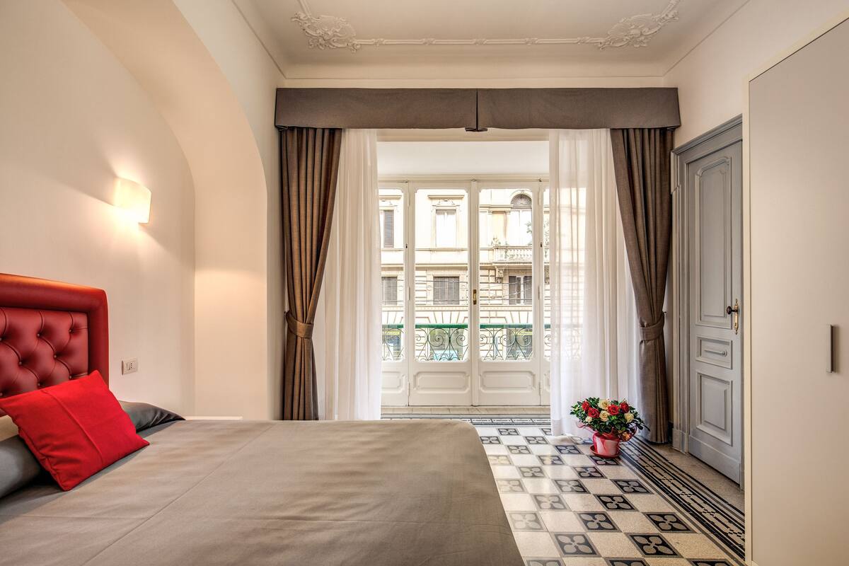 Ein Hotelzimmer mit einem großen Bett, einem roten Kissen, einer Zimmerpflanze und Blick auf einen Balkon mit Geländer.