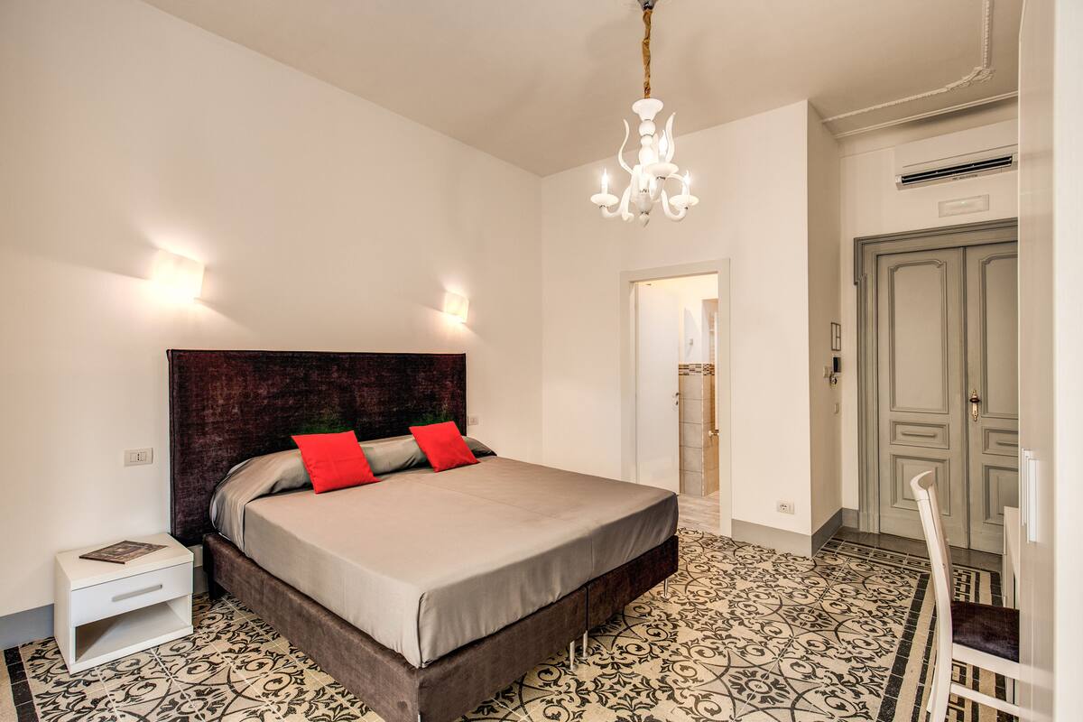 Ein Schlafzimmer mit einem großen Bett, einem Nachttisch, einem Sessel, einer Chandelieber und einer Tür.