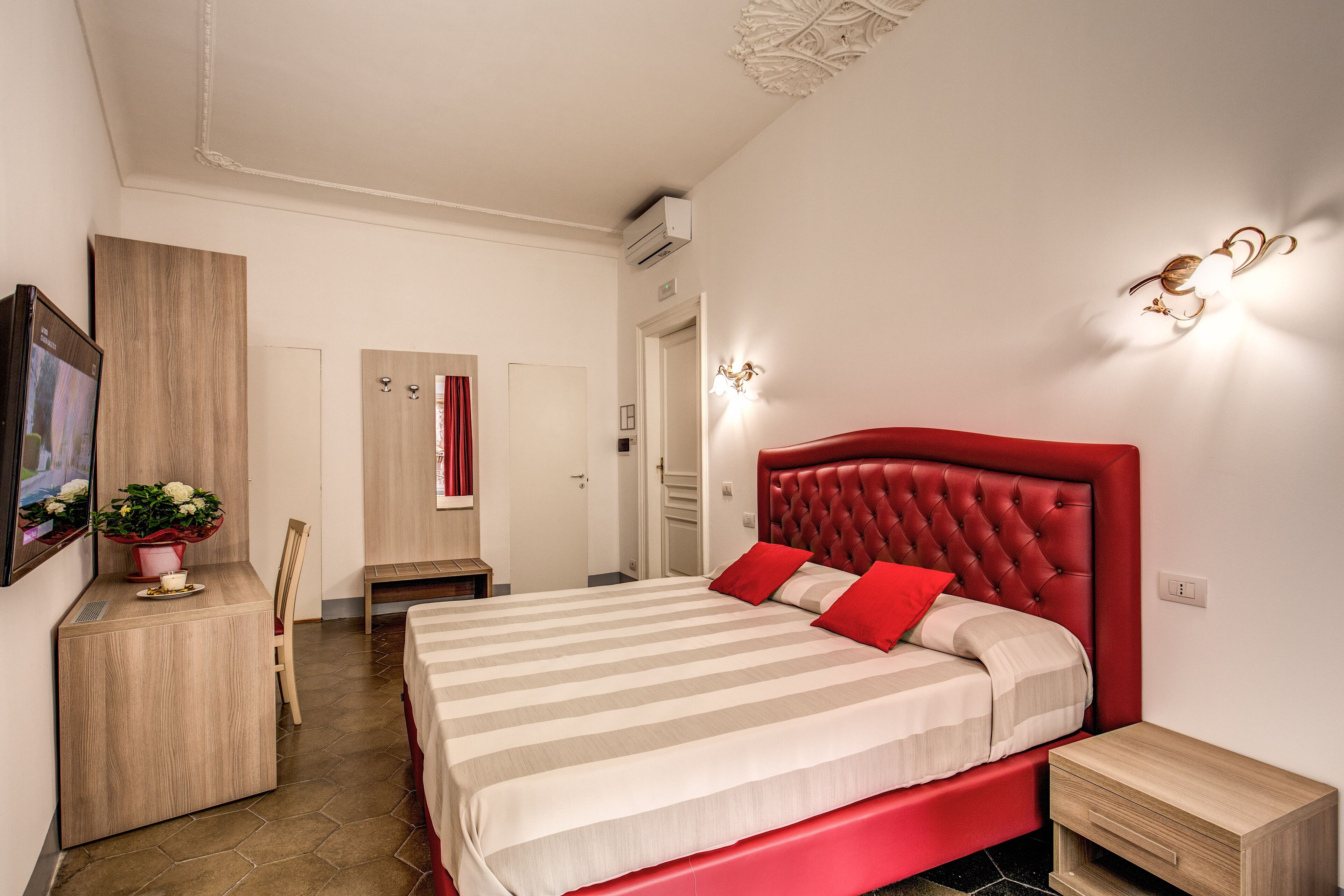 A hotel room with a large bed, a bedside table, a desk with a chair, and a potted plant.