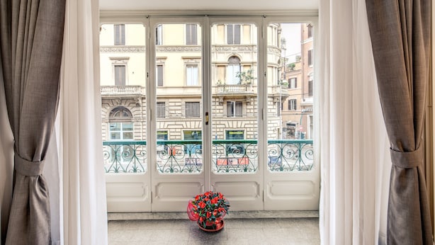 A view from a balcony with a potted plant, a white door, and a building with balconies and railings in the background. - Feronia Guest House (Rome)