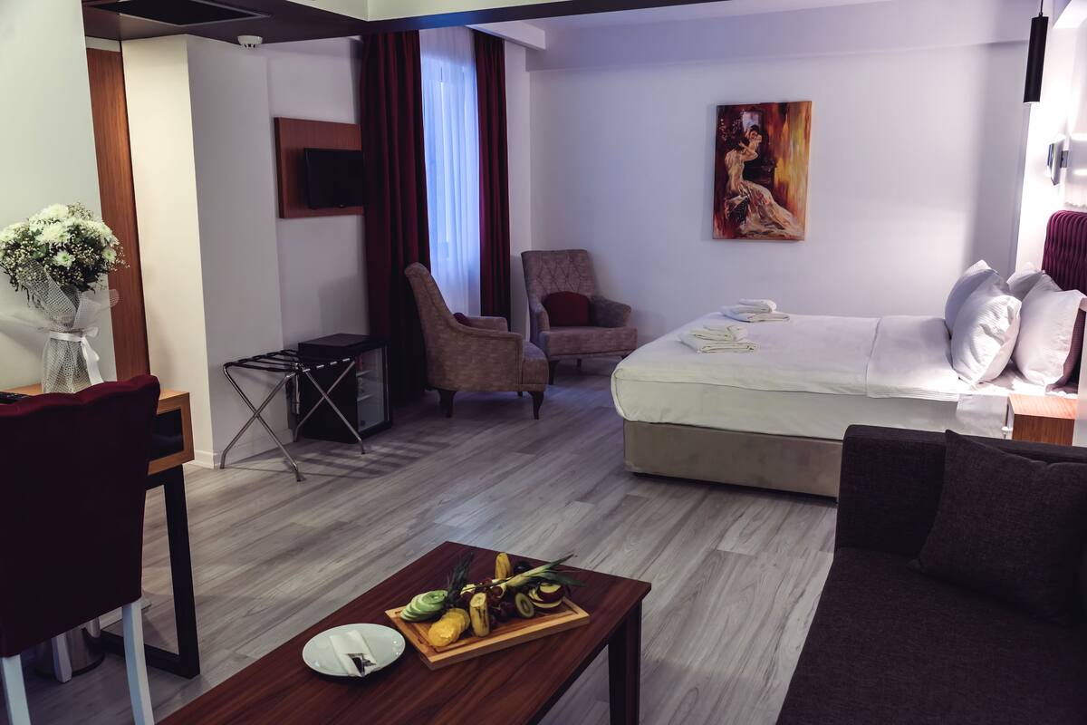 A modern hotel room with a bed, a sofa, a dining table with fruit, a TV, and a painting on the wall.