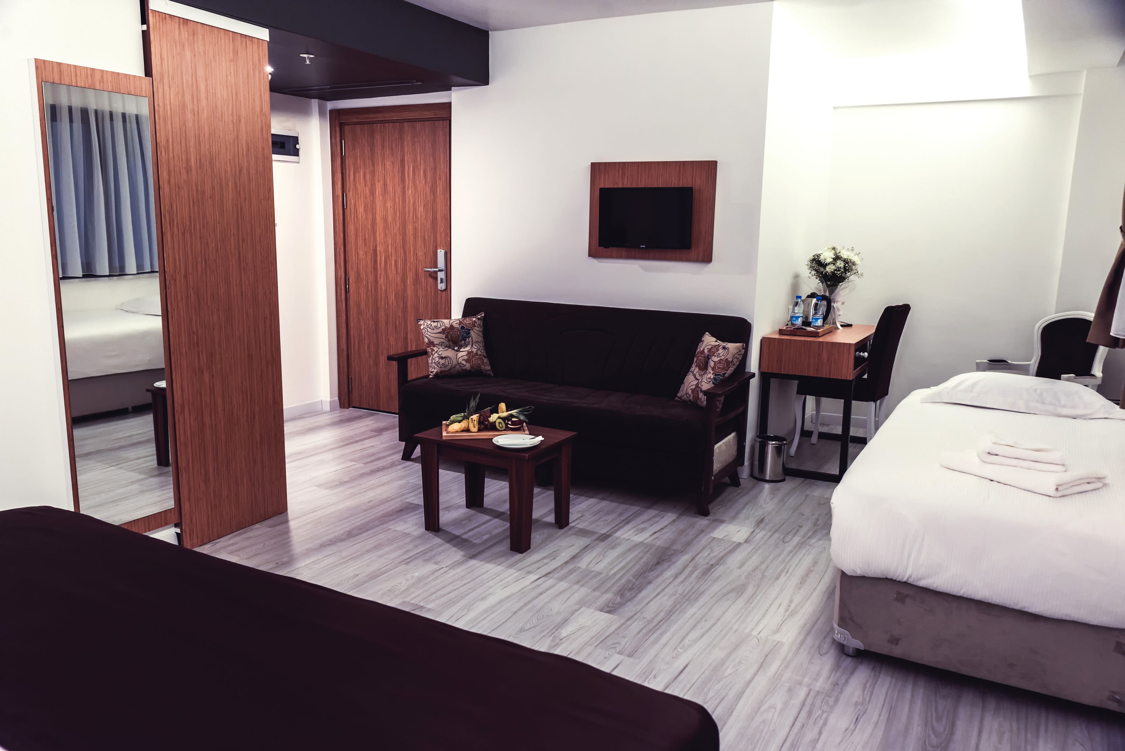 A hotel room with a bed, a sofa, a desk with a computer, a TV, and a small table with fruits.