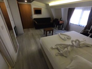Una habitación de hotel con cama, sofá, una mesita, televisor y ventana con cortinas.