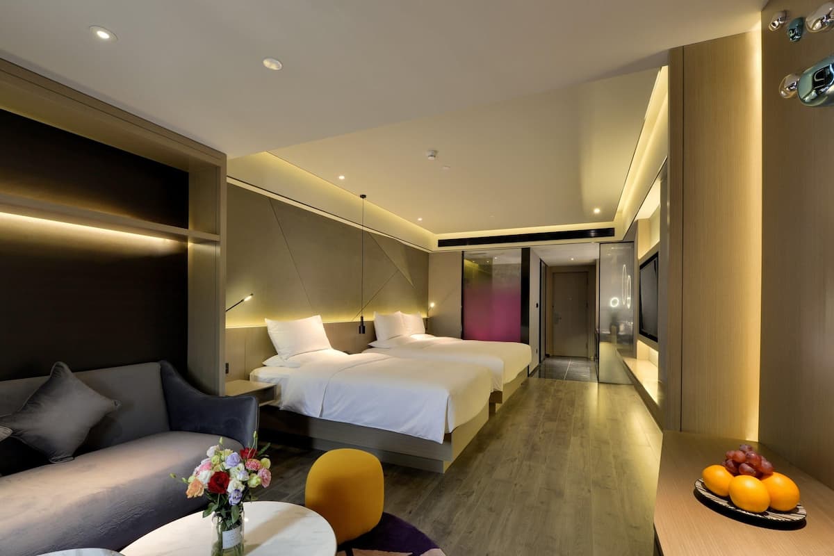 Een moderne hotelkamer met een bank, twee bedden, een klein tafeltje met een bloevas en een schaal fruit.