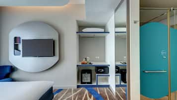 Una habitación de hotel moderna con una cama, un televisor montado en la pared, un escritorio con una computadora y un baño con puerta de vidrio.