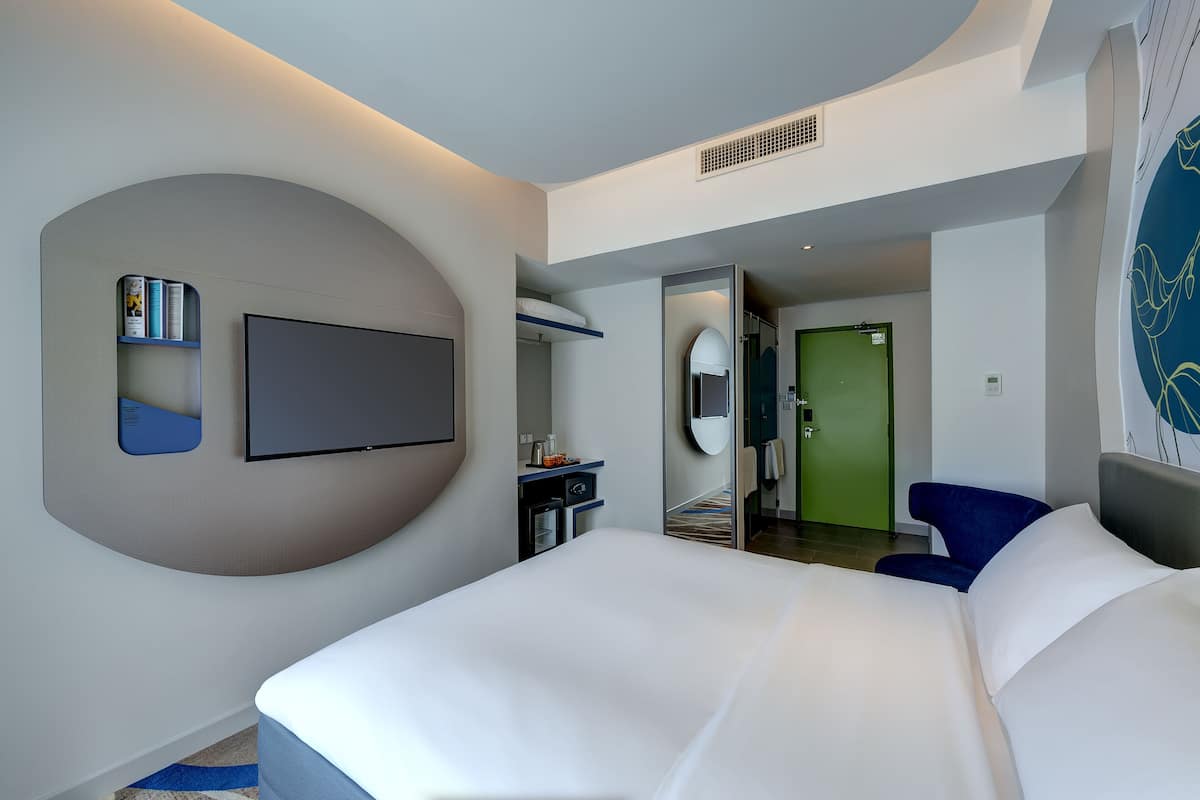 Een moderne hotelkamer met een groot bed, een flatscreen televisie aan de muur, een klein bureau met stoel, en een groene deur die naar een andere kamer leidt.