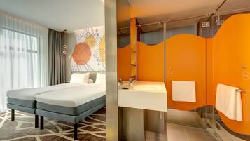 Habitación de hotel con cama doble, ventana grande con cortinas translúcidas, baño moderno con pared de acento naranja y cabina de ducha de vidrio.