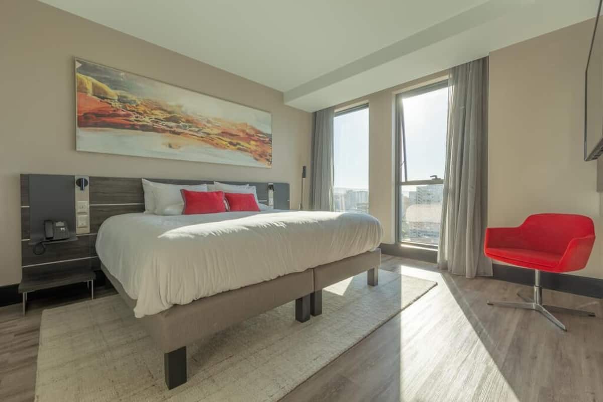 A modern hotel room with a large bed, a red chair, a painting on the wall, and a city view.