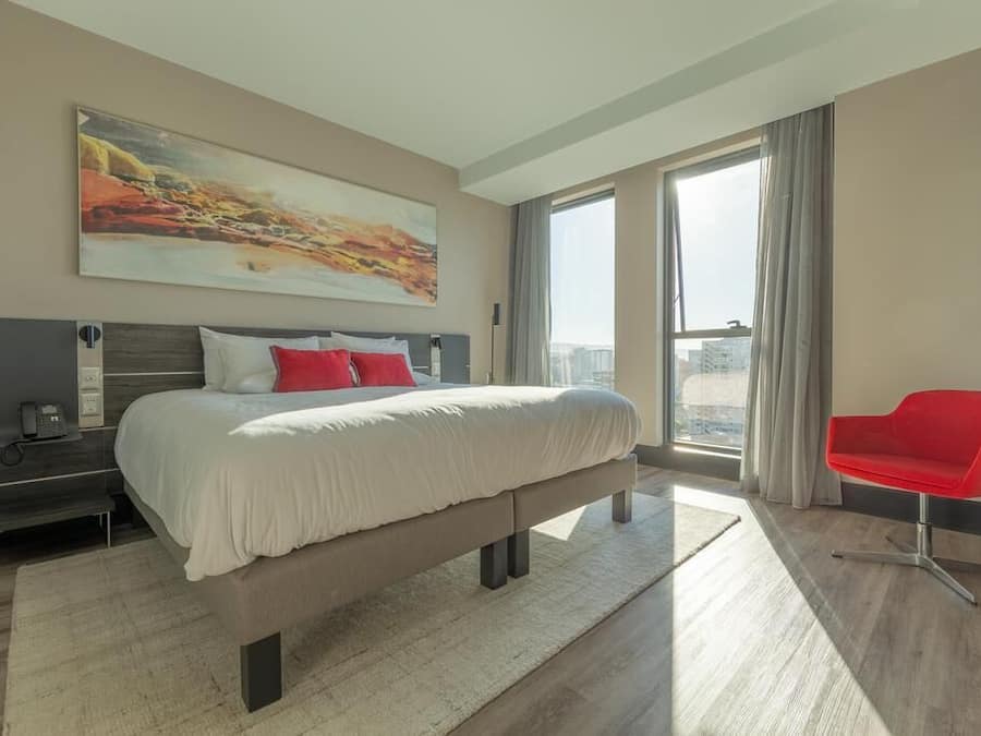 Una camera d'hotel moderna con un letto grande, una poltrona rossa, un quadro appeso e vista sulla città.