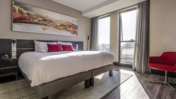 Quarto de hotel moderno com cama grande, uma cadeira vermelha, um quadro na parede e vista para a cidade.