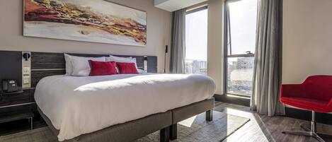 Een moderne hotelkamer met een groot bed, een rode stoel, een schilderij aan de muur en uitzicht over de stad.