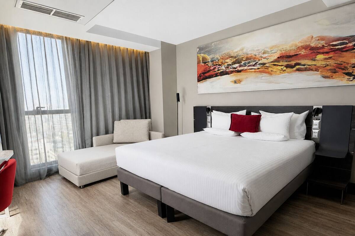 A modern hotel room with a large bed, a sofa, a red chair, and a painting on the wall.
