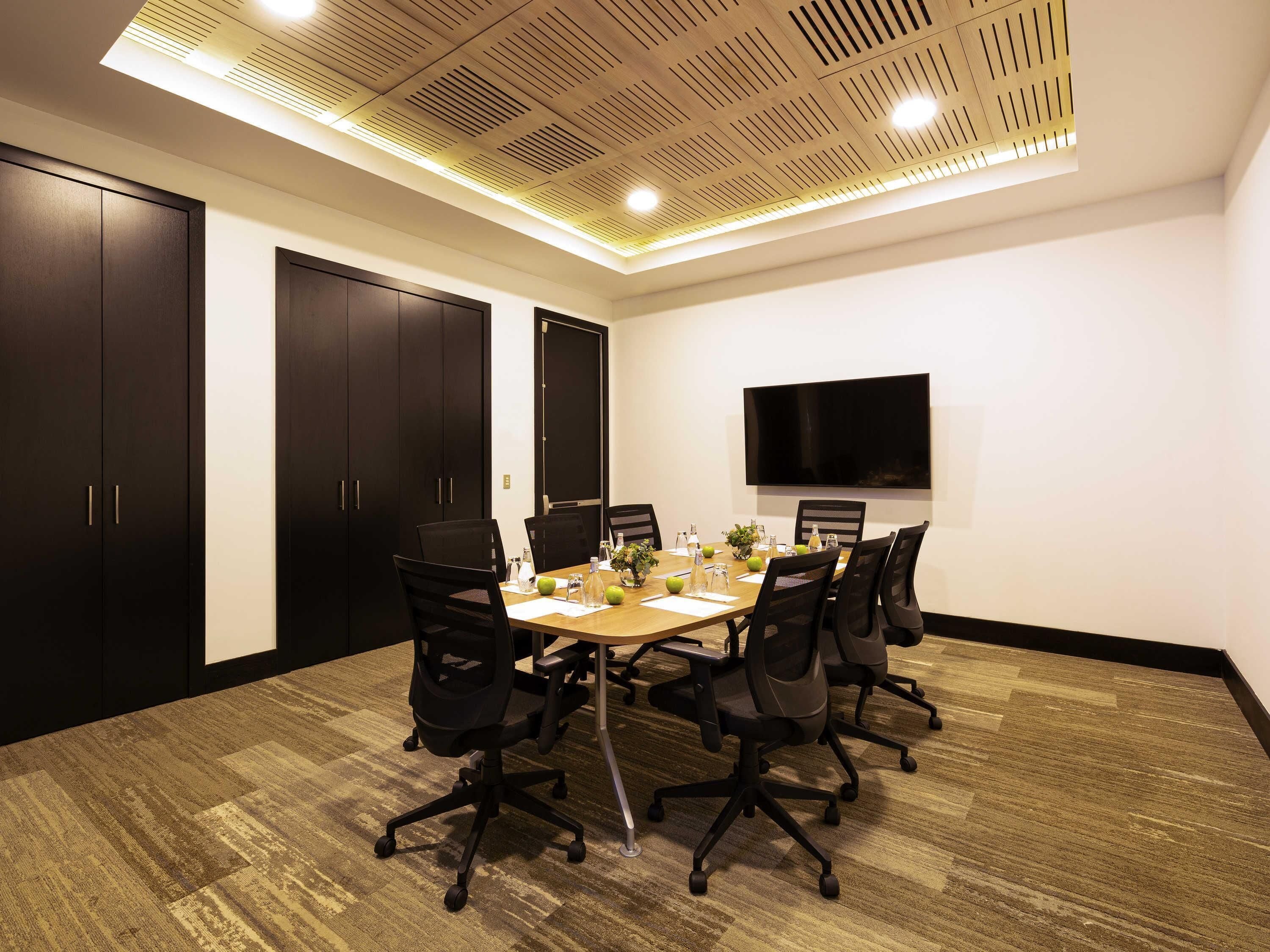 A conference room with a large table, black chairs, a flat-screen TV, and a ceiling with recessed lighting.