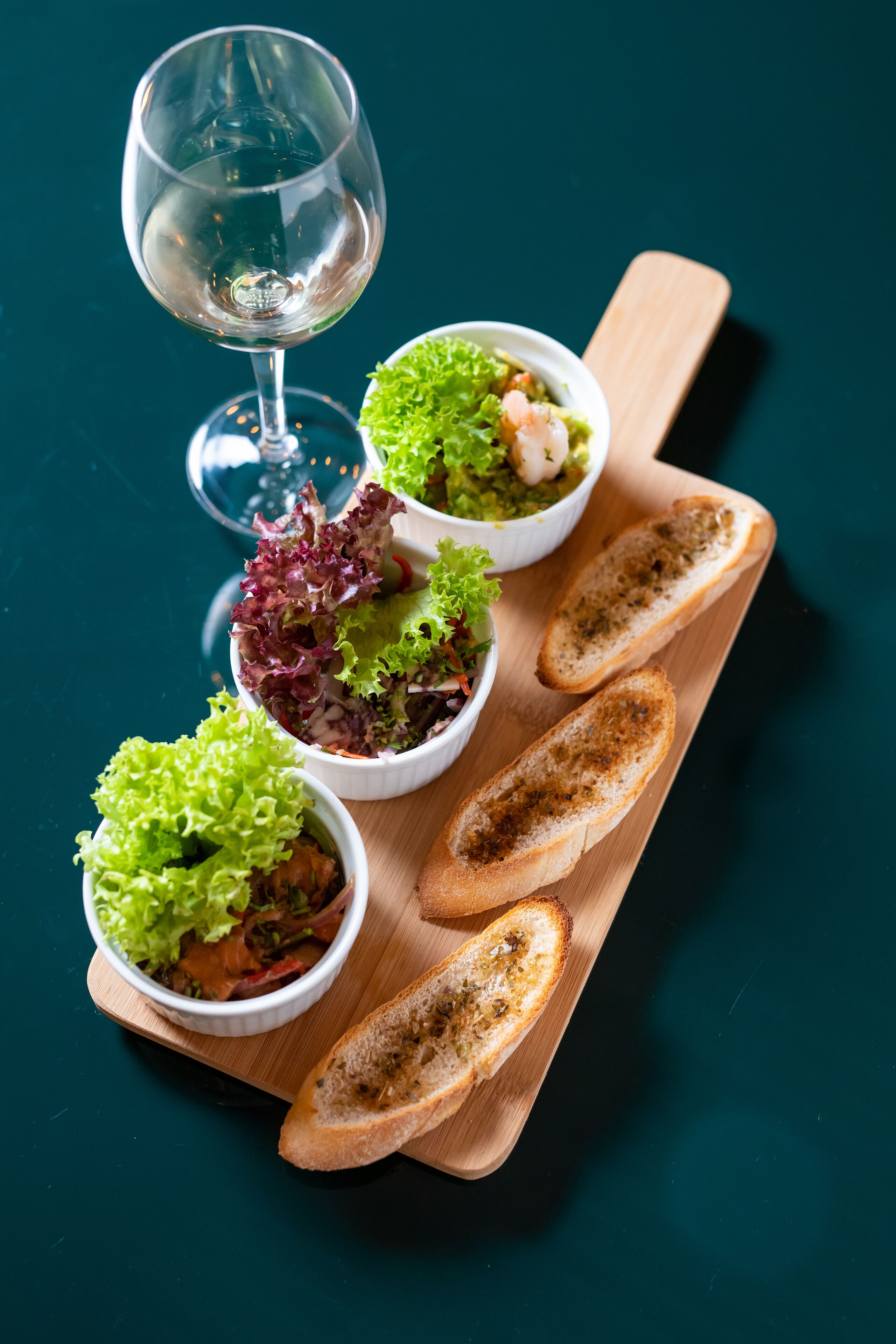 Una tabla de madera con cuatro ensaladas en cuencos y dos rebanadas de pan tostado, acompañada de una copa de vino.