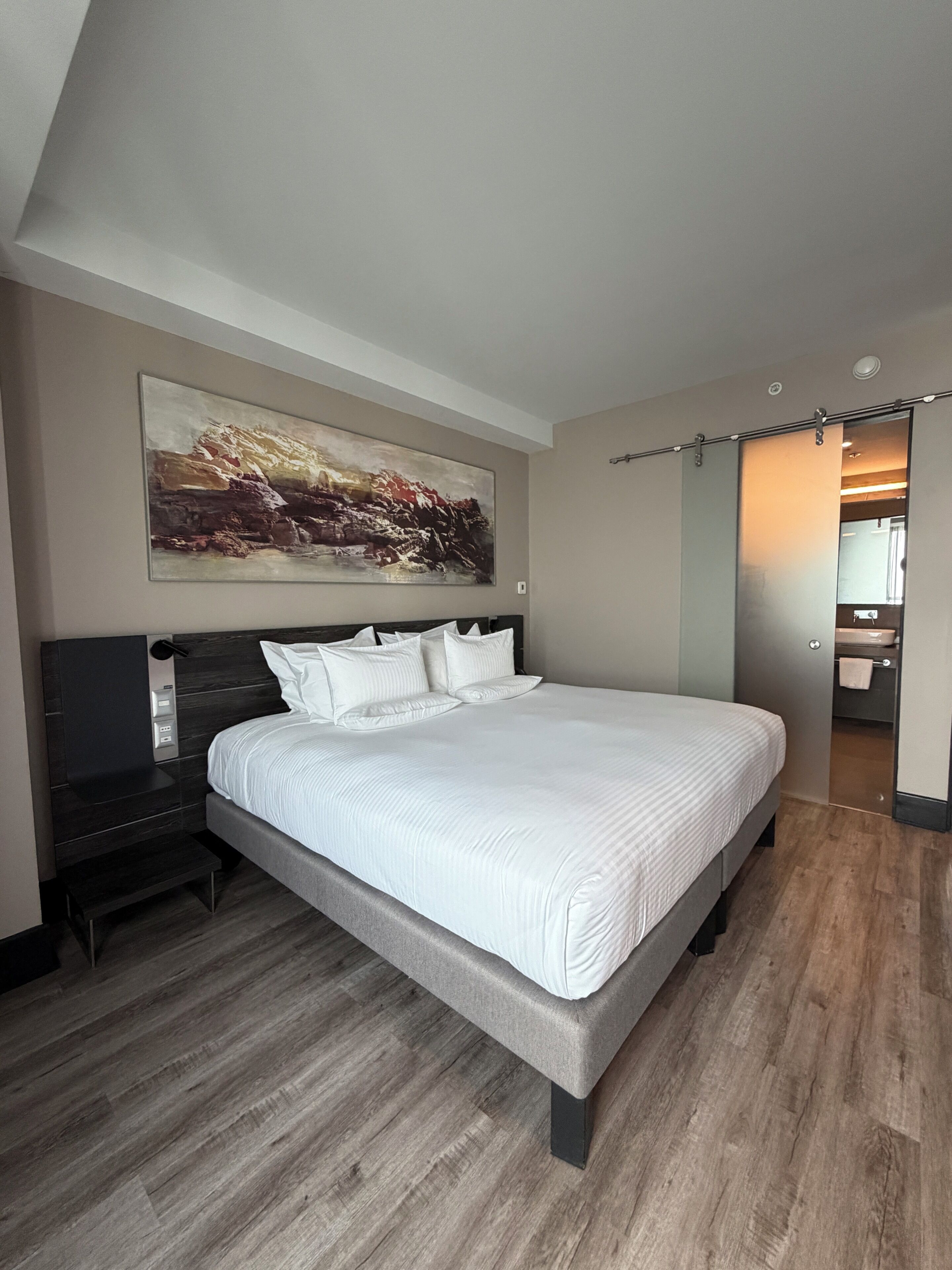 Un dormitorio con cama, mesita de noche, un cuadro y un baño visible a través de una puerta corredera.