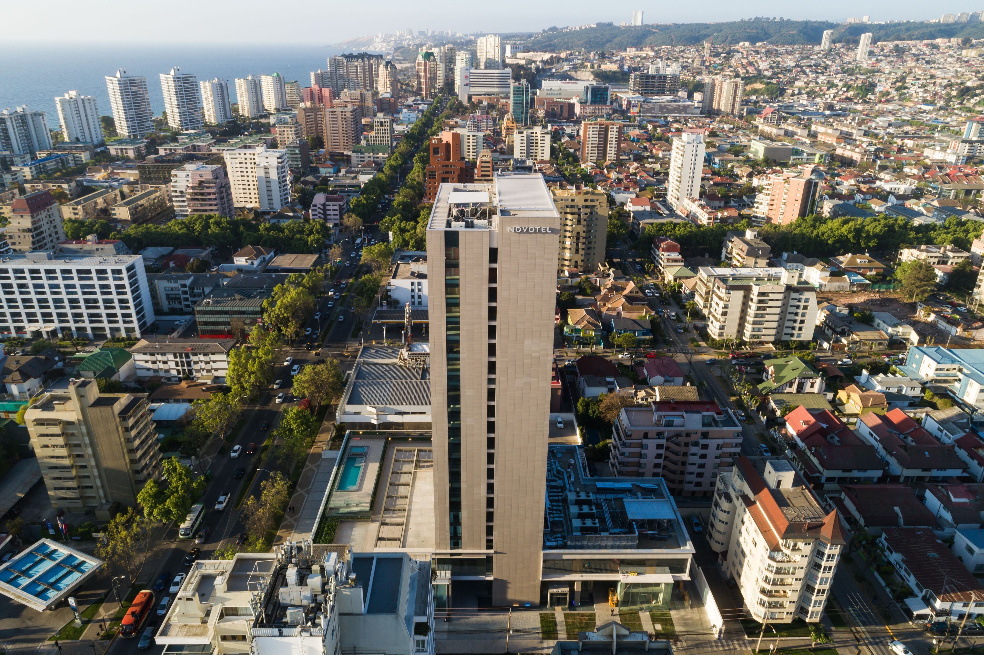 Un hôtel de grande hauteur au design moderne, situé au cœur d’un quartier urbain dense, entouré de différents bâtiments et offrant une vue dégagée sur la mer en arrière-plan.