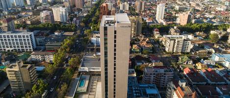 A high-rise hotel with a modern design, surrounded by a dense urban area with various buildings and a clear view of the sea in the background.