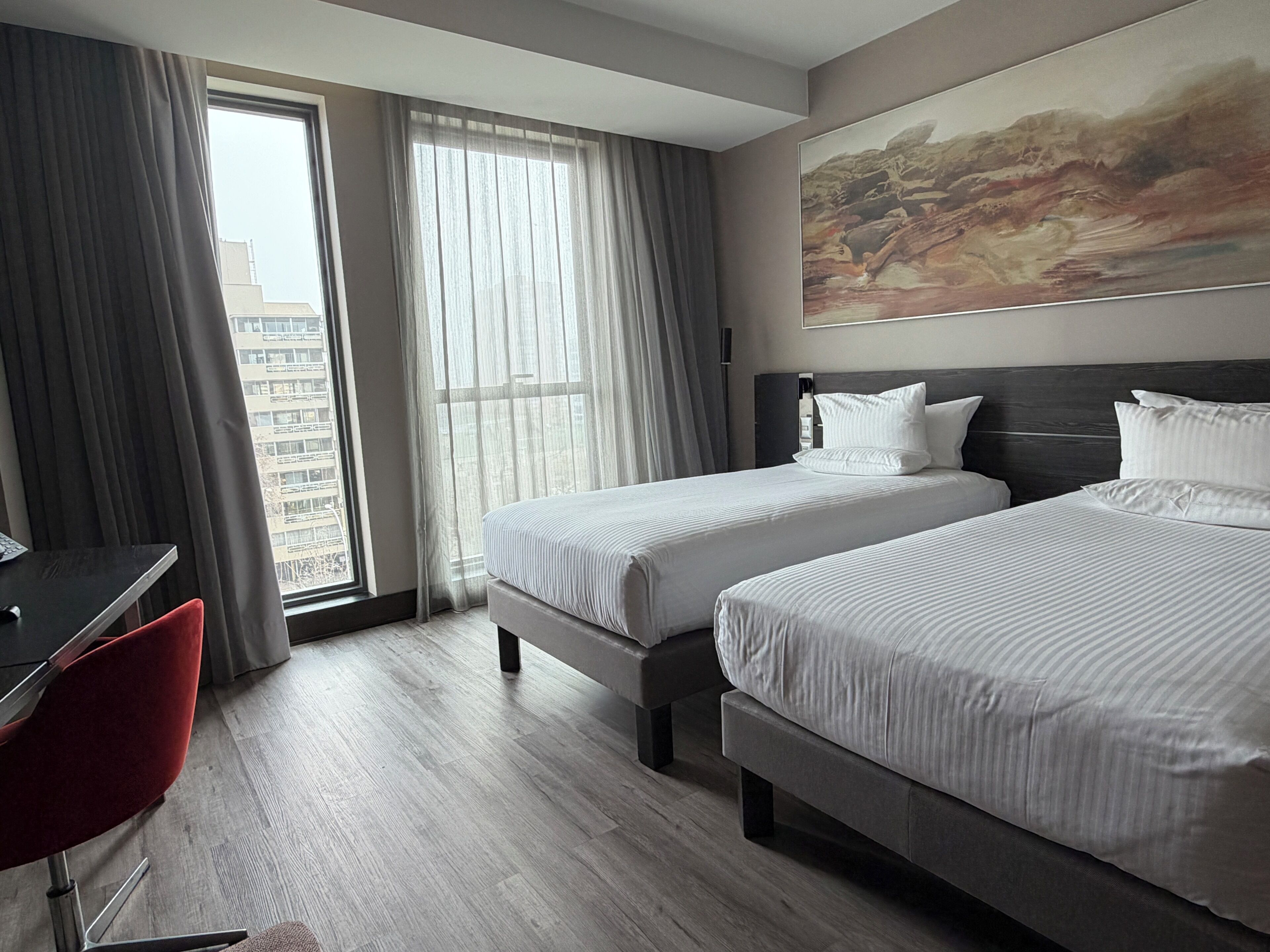 Ein Hotelzimmer mit zwei Betten, einem Schreibtisch, einem Stuhl und einem großen Fenster mit Vorhängen.