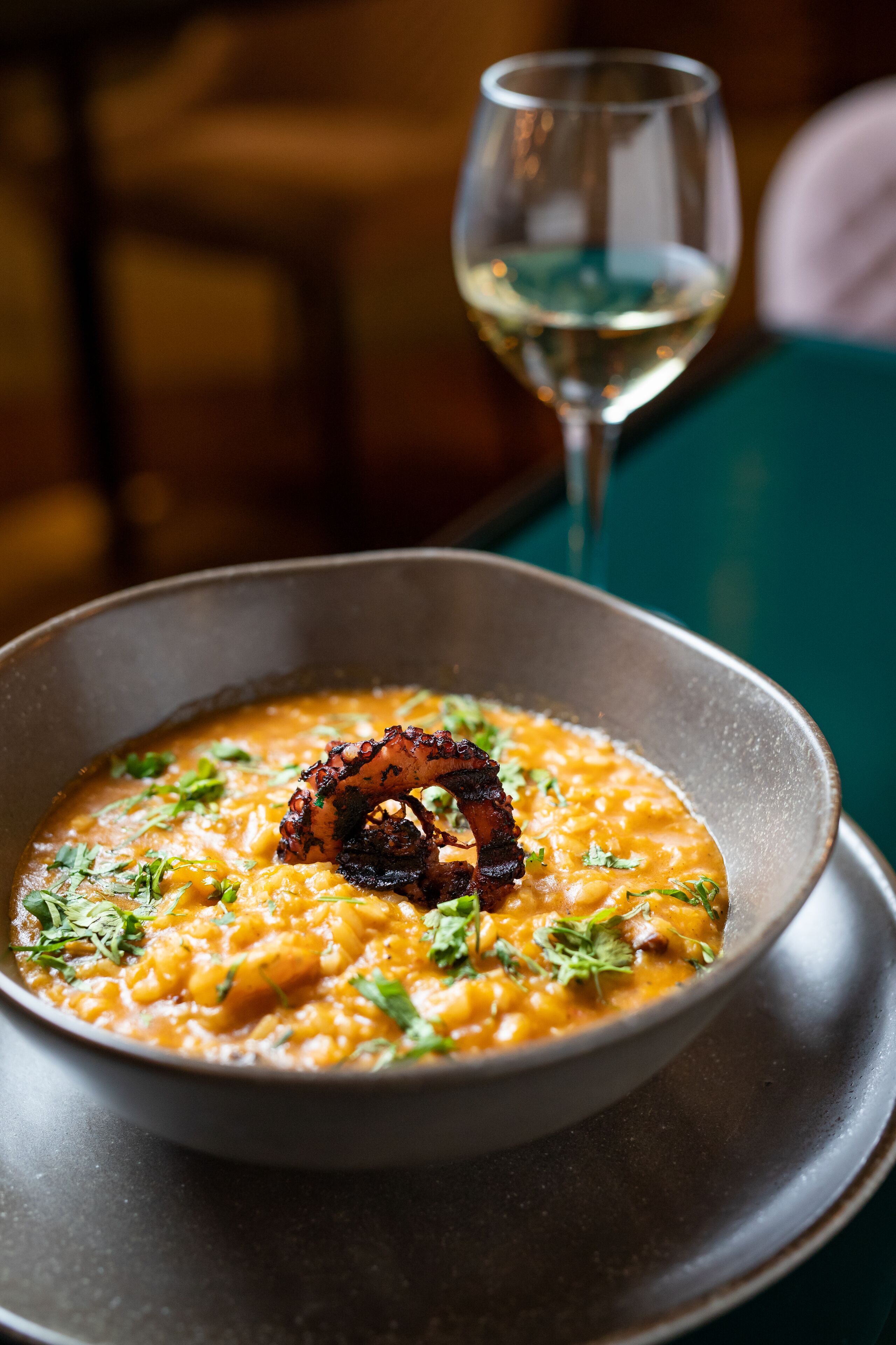 A bowl of soup with a garnish, a glass of white wine, and a dark table.