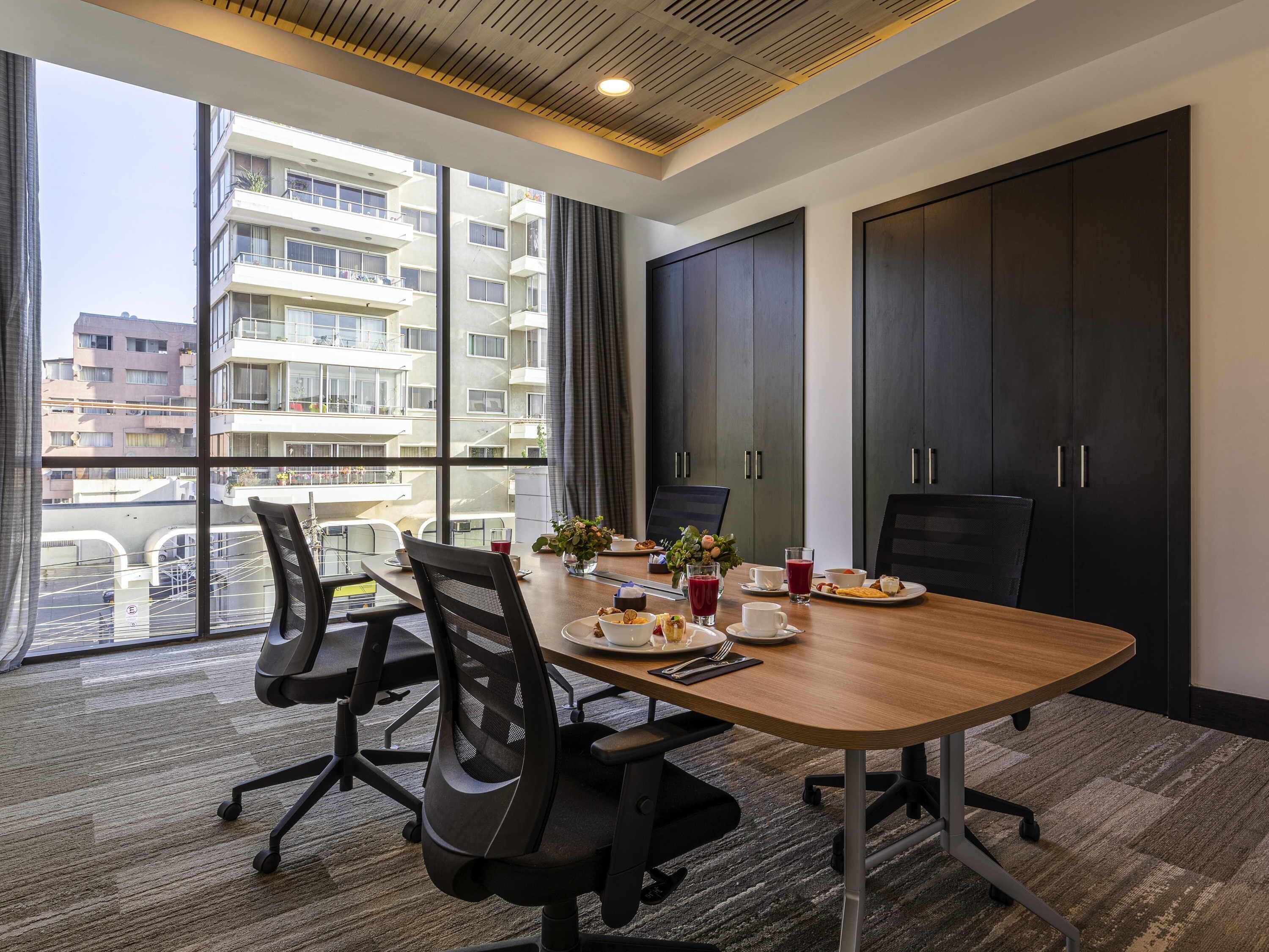 Un salón de conferencias con una gran mesa, sillas, una computadora portátil y vistas a un edificio residencial.
