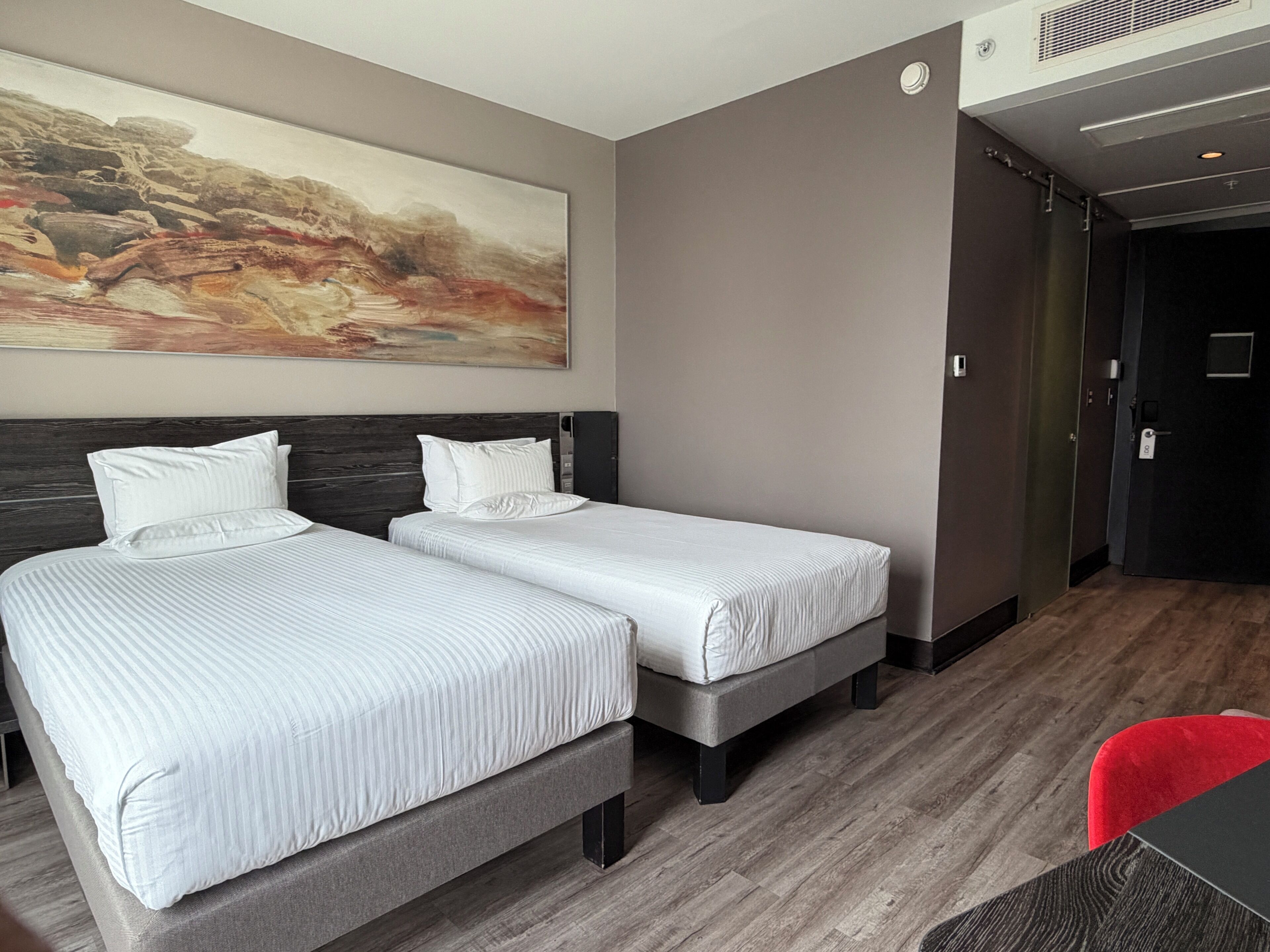 A hotel room with two beds, a large abstract painting on the wall, a dark door, and a red chair.