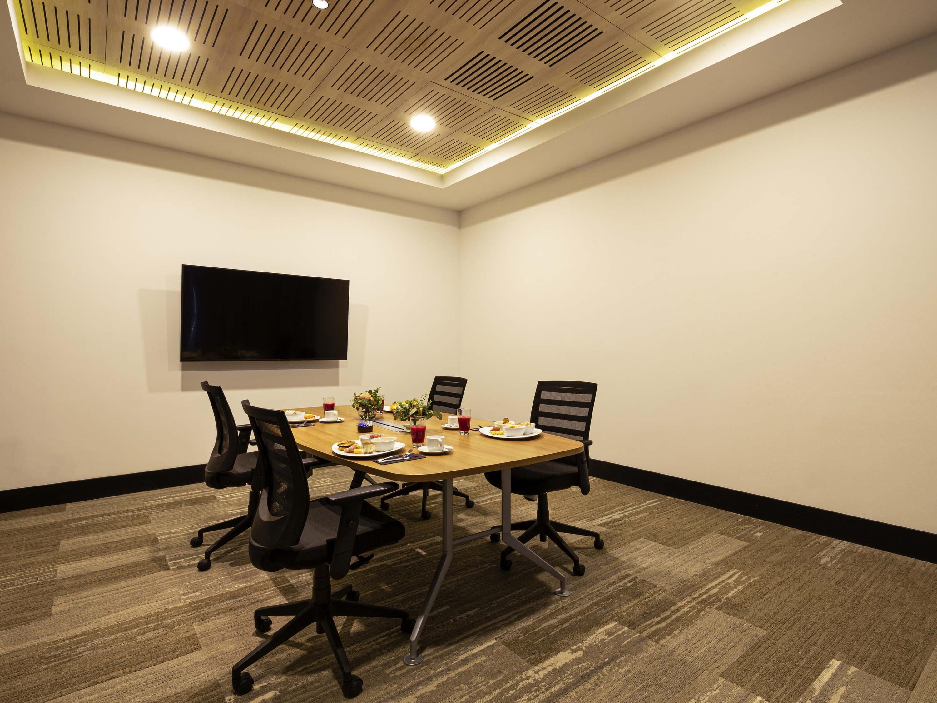 A conference room with a long table, four chairs, a flat-screen TV, and a ceiling with recessed lighting.