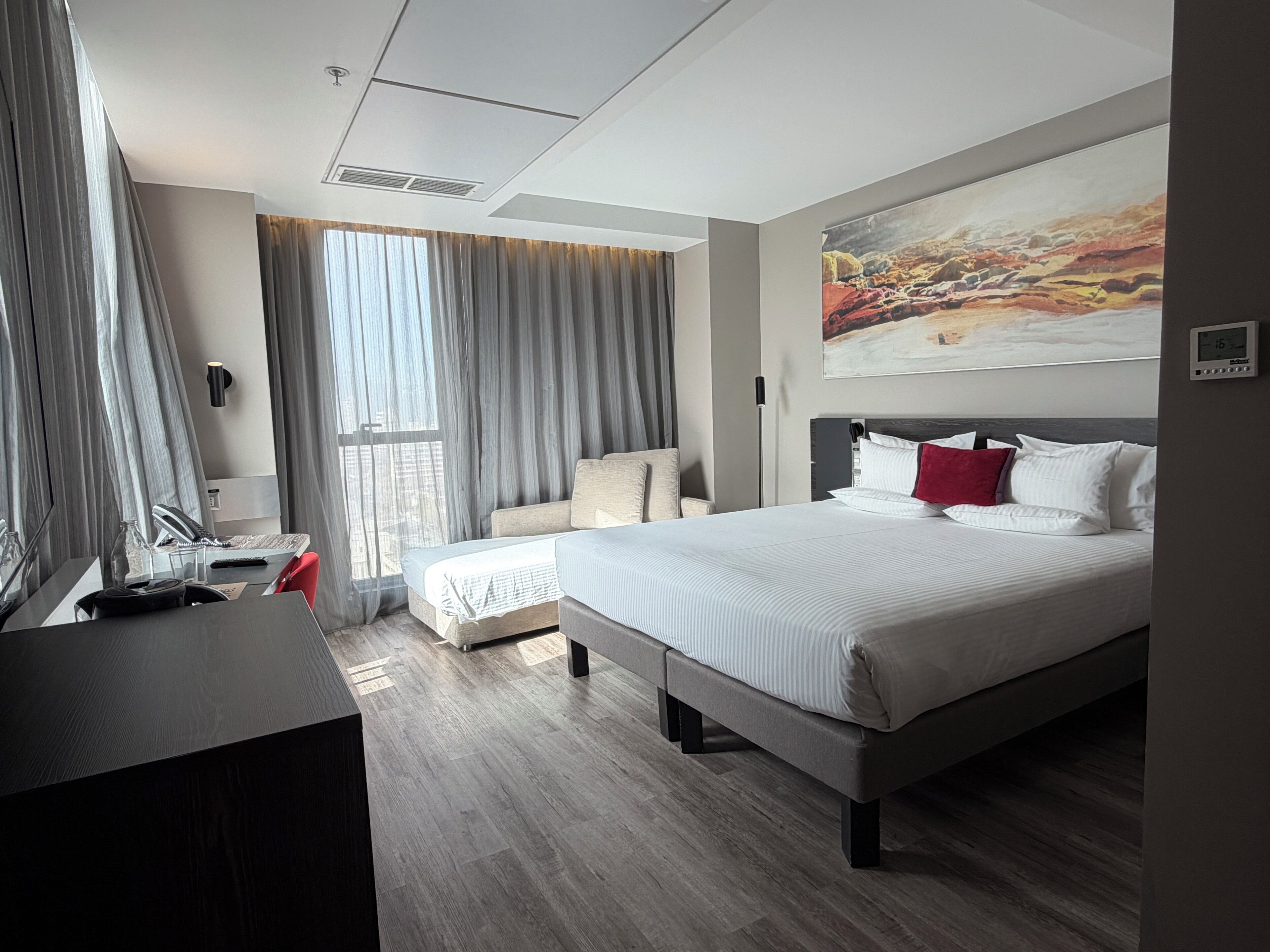 A modern hotel room with a large bed, a desk, a chair, and a painting on the wall.