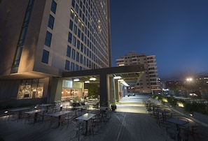 An outdoor dining area with tables and chairs, a covered seating area, and a view of a cityscape at dusk.