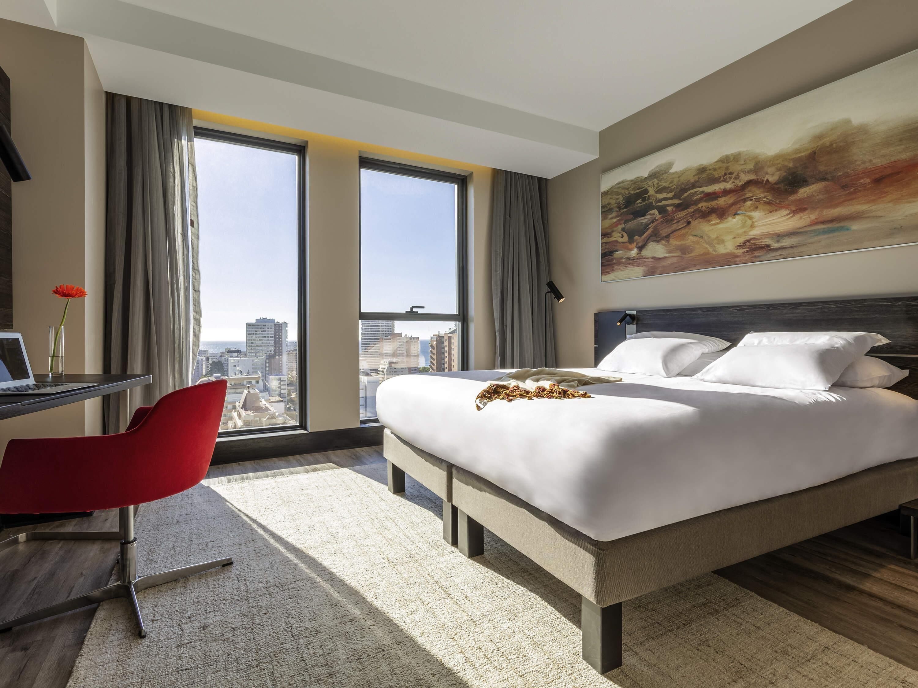 A hotel room with a large bed, a desk with a laptop, a red chair, and a city view.