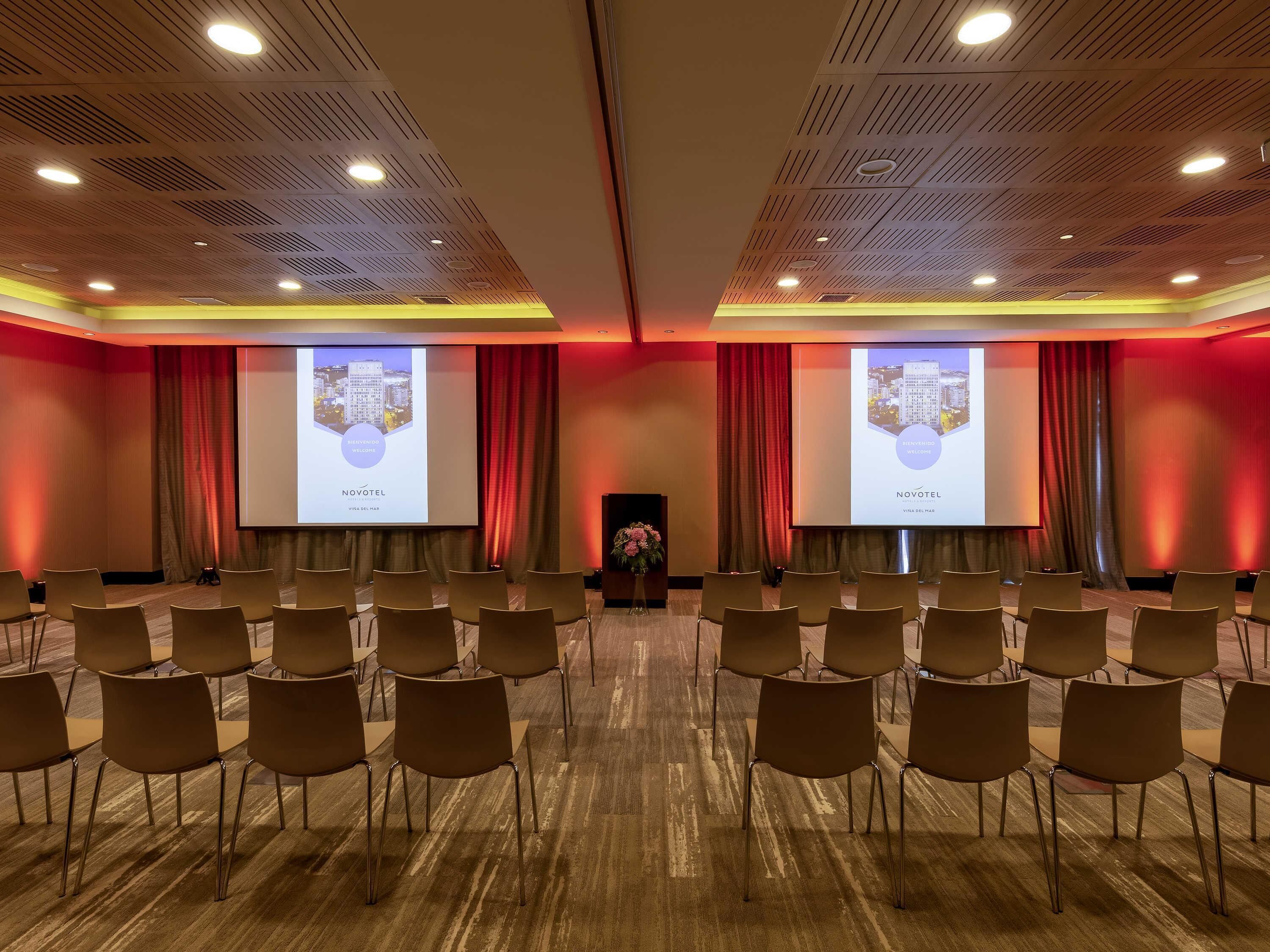 A conference room with rows of chairs, a podium, and two large projection screens displaying a welcome message.