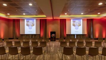 Un salón de conferencias con filas de sillas, un atril y dos grandes pantallas de proyección que muestran un mensaje de bienvenida.