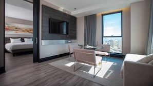 Een moderne hotelkamer met een bed, een aan de muur bevestigde televisie, een zithoek met een tafel en stoelen, en een groot raam met uitzicht over de stad.
