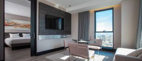 Una habitación de hotel moderna con una cama, un televisor montado en la pared, una zona de comedor con una mesa y sillas, y un ventanal con vistas a la ciudad.