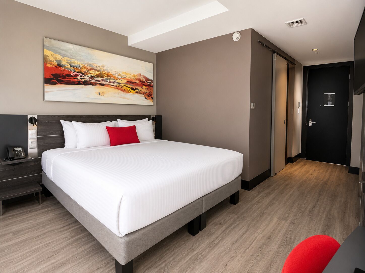 A modern hotel room with a large bed, a bedside table with a phone, a painting on the wall, and a door to the right.