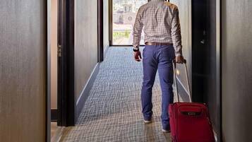Un hombre camina por un pasillo con una maleta roja, hacia una puerta que da a vistas de edificios.