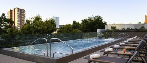 Une piscine sur le toit offrant une vue dégagée sur les bâtiments environnants, une terrasse en bois et des chaises longues avec des serviettes.