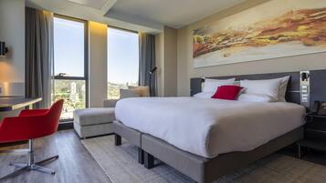 Una habitación de hotel moderna con una cama grande, un escritorio, una silla roja y vistas a la ciudad.