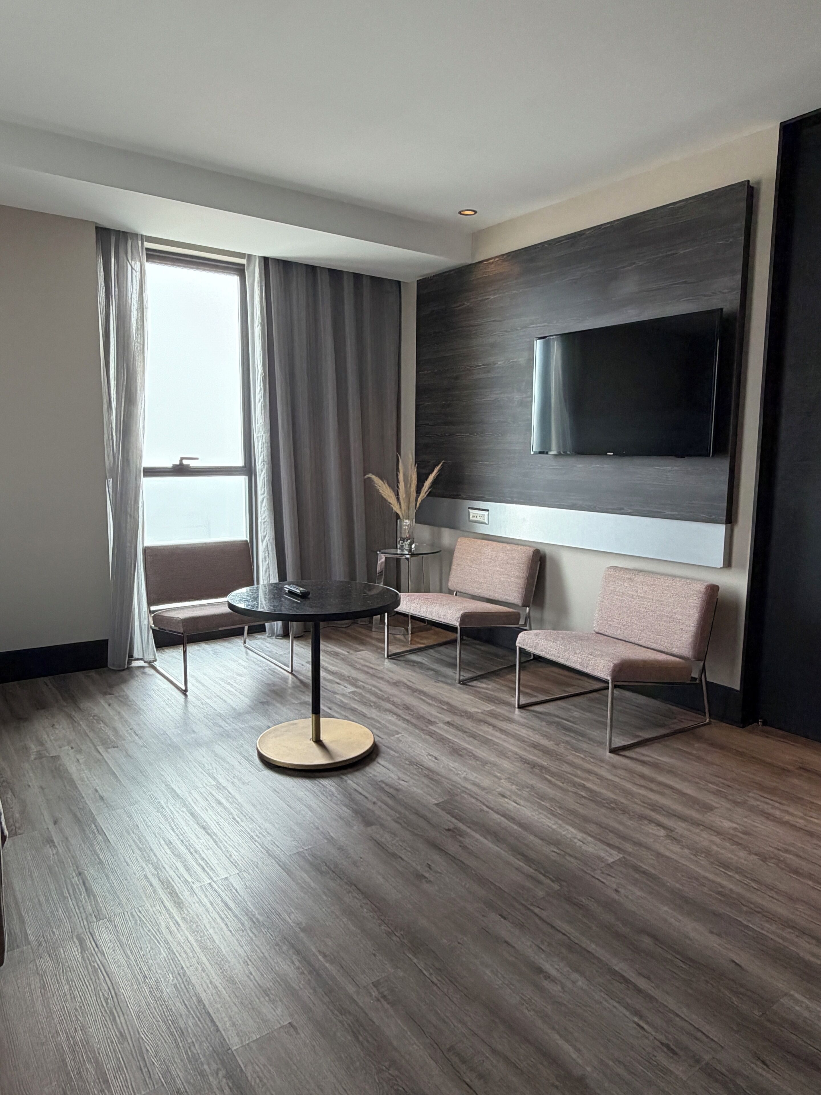 A modern hotel room with a round table, two sofas, a flat-screen TV mounted on the wall, and a large window with curtains.