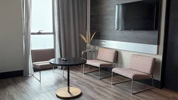 A modern hotel room with a round table, two sofas, a flat-screen TV mounted on the wall, and a large window with curtains.