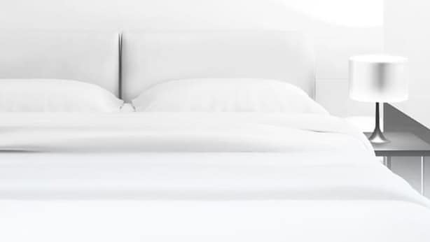 Uma cama com roupa branca, uma mesa de cabeceira com abajur e uma parede lisa.