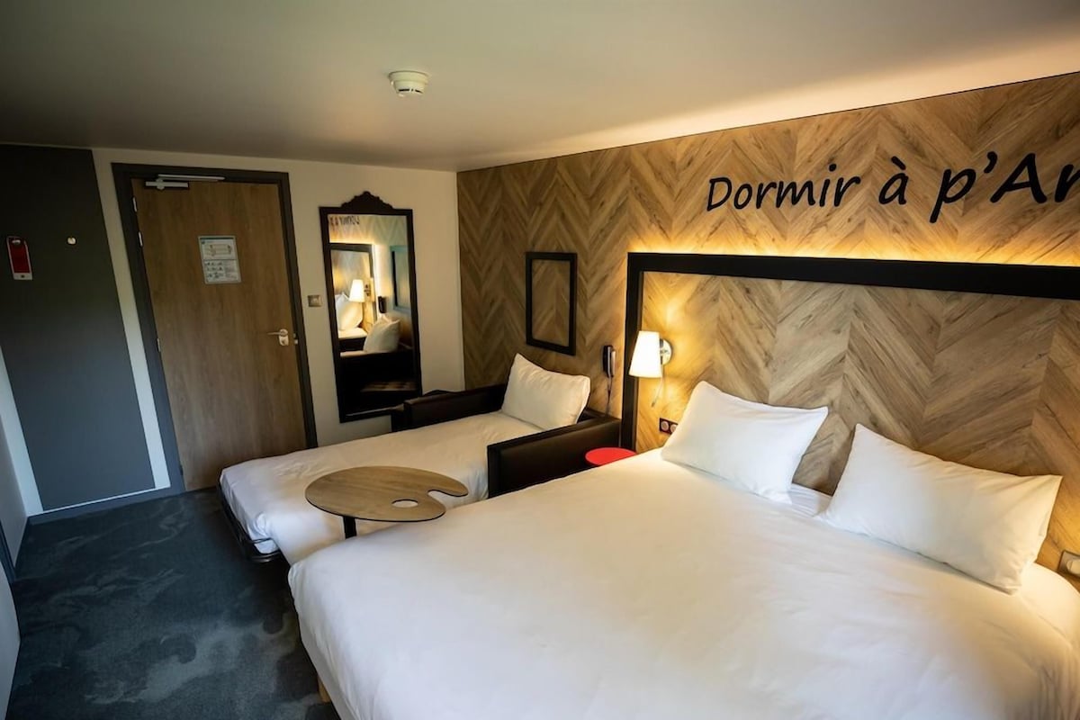 A hotel room with two beds, a wooden headboard, a bedside table, a lamp, and a mirror.