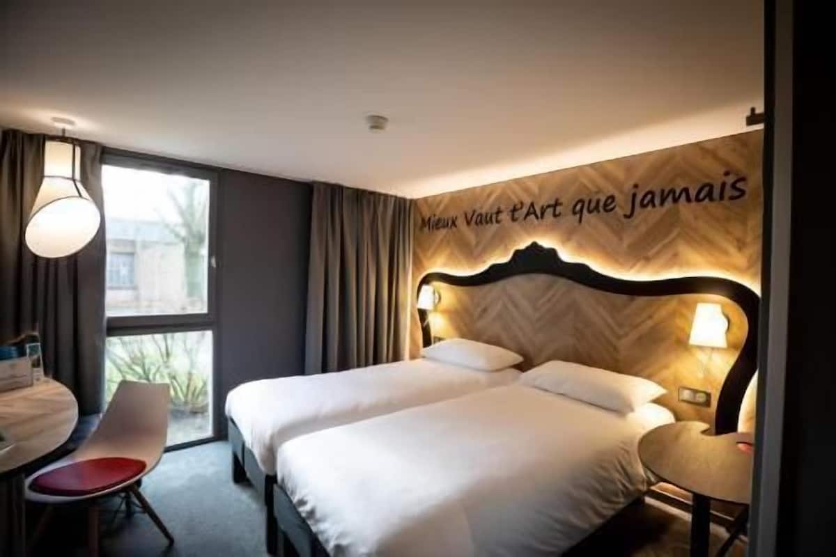 A hotel room with a double bed, a desk with a chair, and a large window.