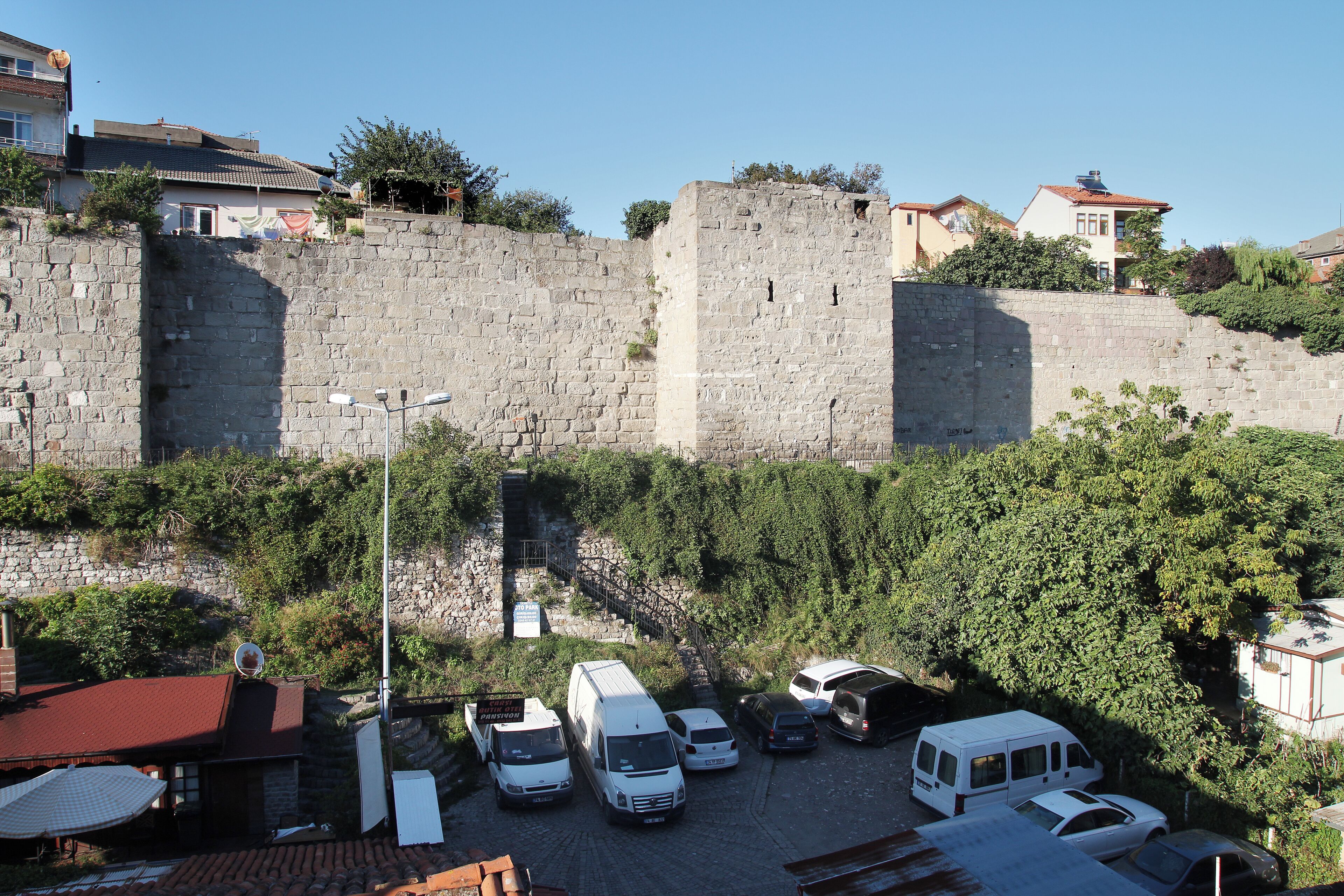 Un estacionamiento con varias camionetas blancas, un muro de piedra y edificios al fondo.