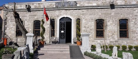 A stone building with the sign "MUZE" above the entrance, featuring a flag, potted plants, and statues.