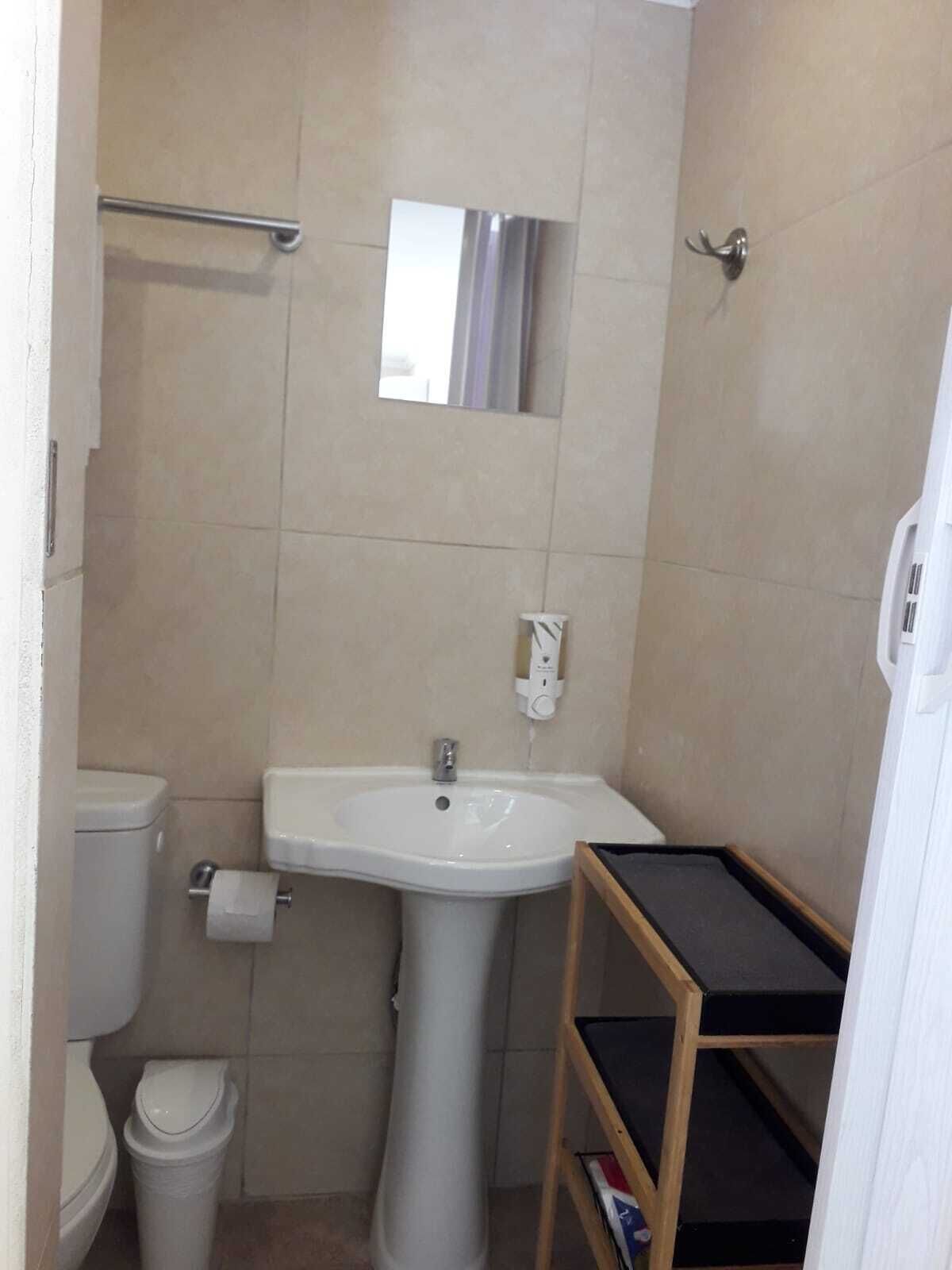 A small bathroom with a white sink, toilet, and trash can. There is a mirror above the sink and a shelf with a black top.